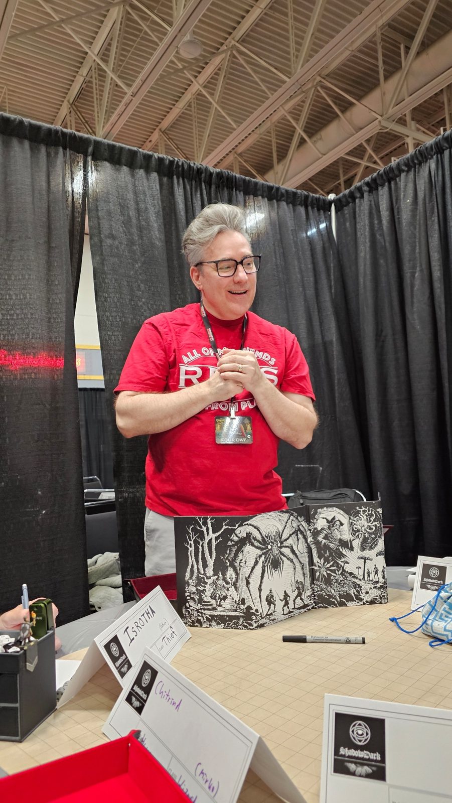 Sean behind a GM screen at a convention, grinning, in a red shirt