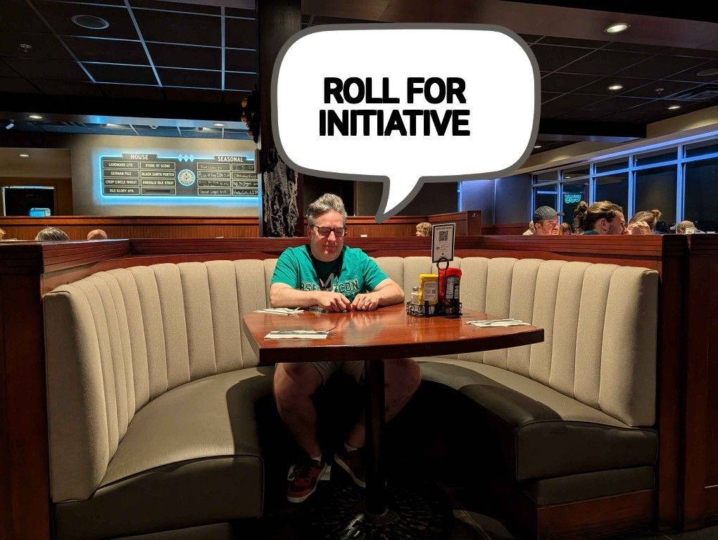 Sean sitting alone in a large restaurant booth with a speech bubble reading ROLL FOR INITIATIVE